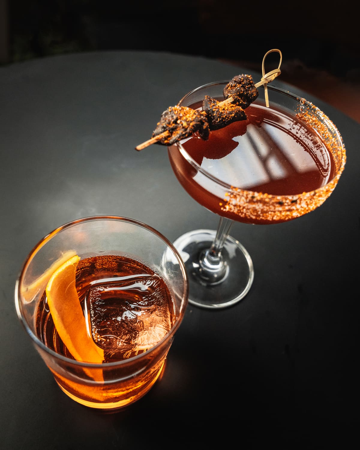 Two cocktails from The Foundry Nagoa's bar: one over a large ice cube with orange peel, one in a coupe with a skewer garnish